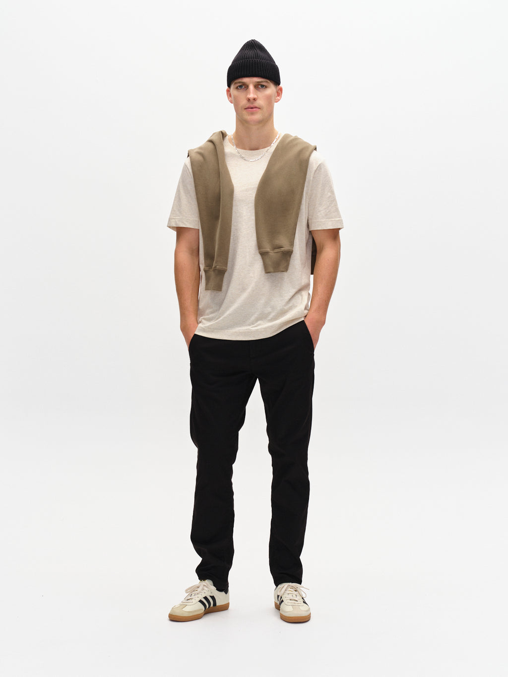 A man in a beanie and casual outfit stands against a plain white backdrop, wearing GABBAs Paul K3280 Dale Slim Chino in black with French pockets. He pairs them with a beige T-shirt, branded buttoned sweater draped over his shoulders, and stylish white sneakers with black stripes.