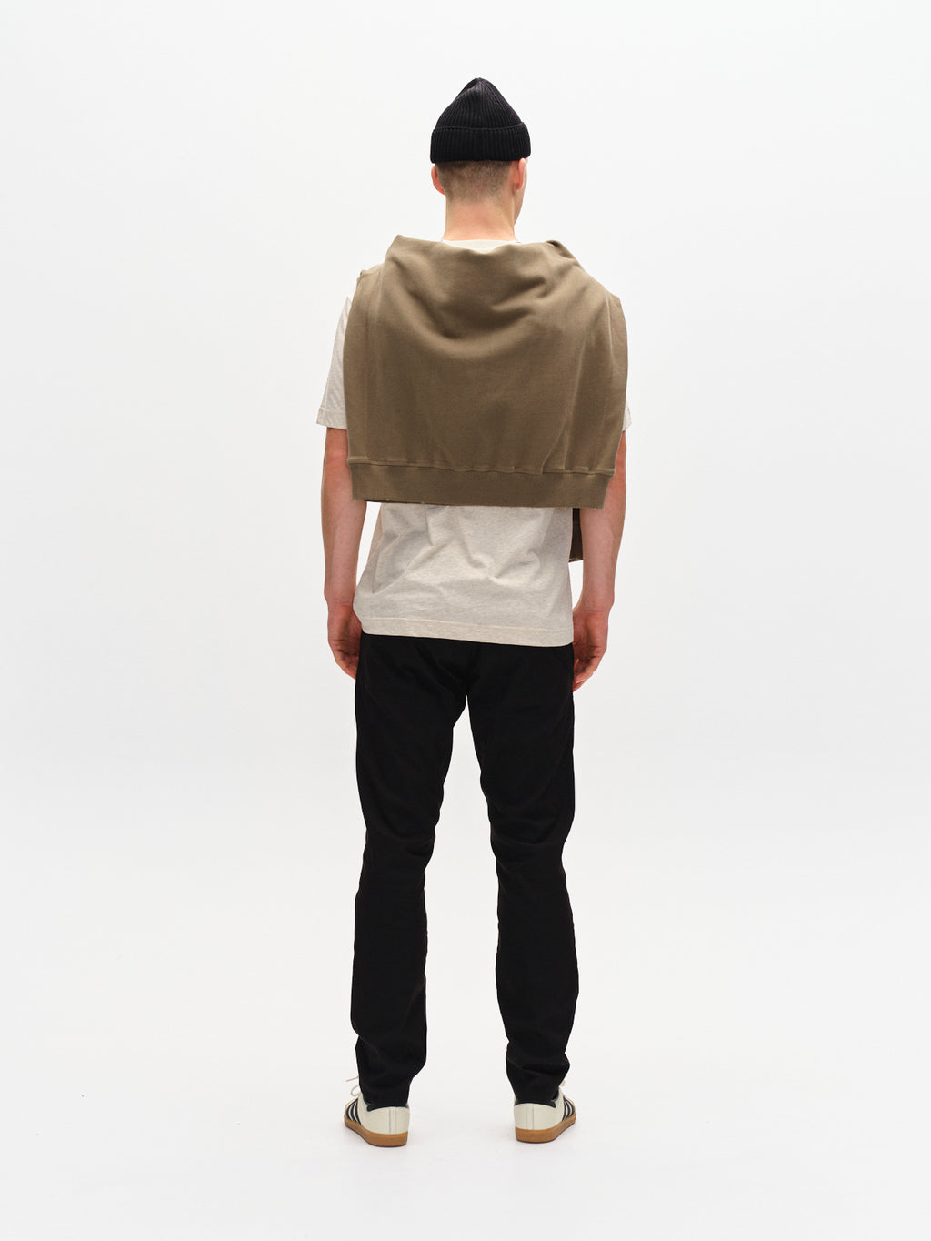 A person stands facing away, wearing a white t-shirt, Paul K3280 Dale Chino - Black pants by GABBA with French pockets, sneakers, and a beanie. A brown sweater is draped over their shoulders against a plain white background.