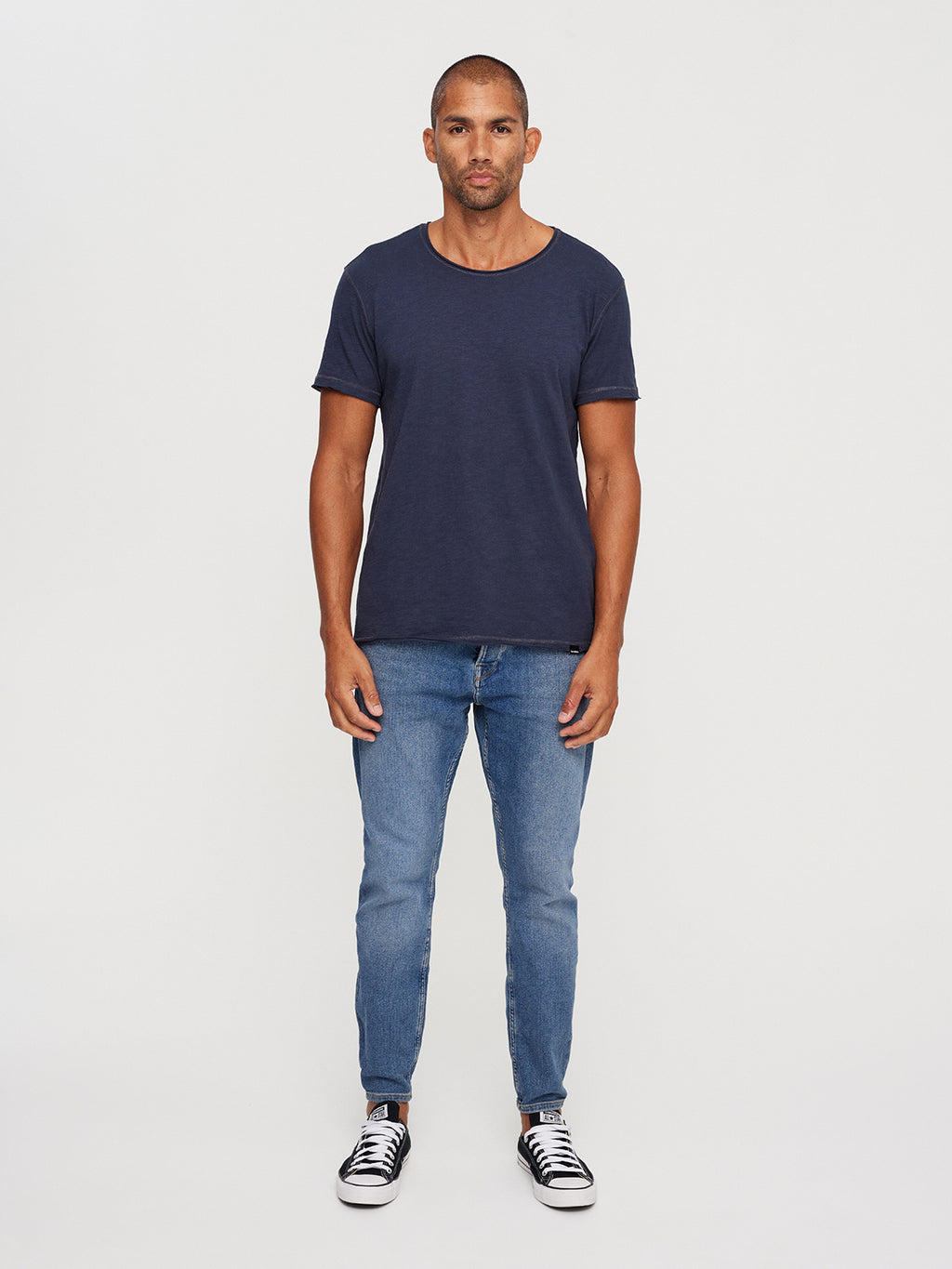 A man models GABBA’s Konrad Straight Slub Tee in navy, paired with blue jeans and black sneakers with white soles. His short hair and neutral expression enhance the casual vibe, with arms relaxed by his sides against a plain white backdrop.