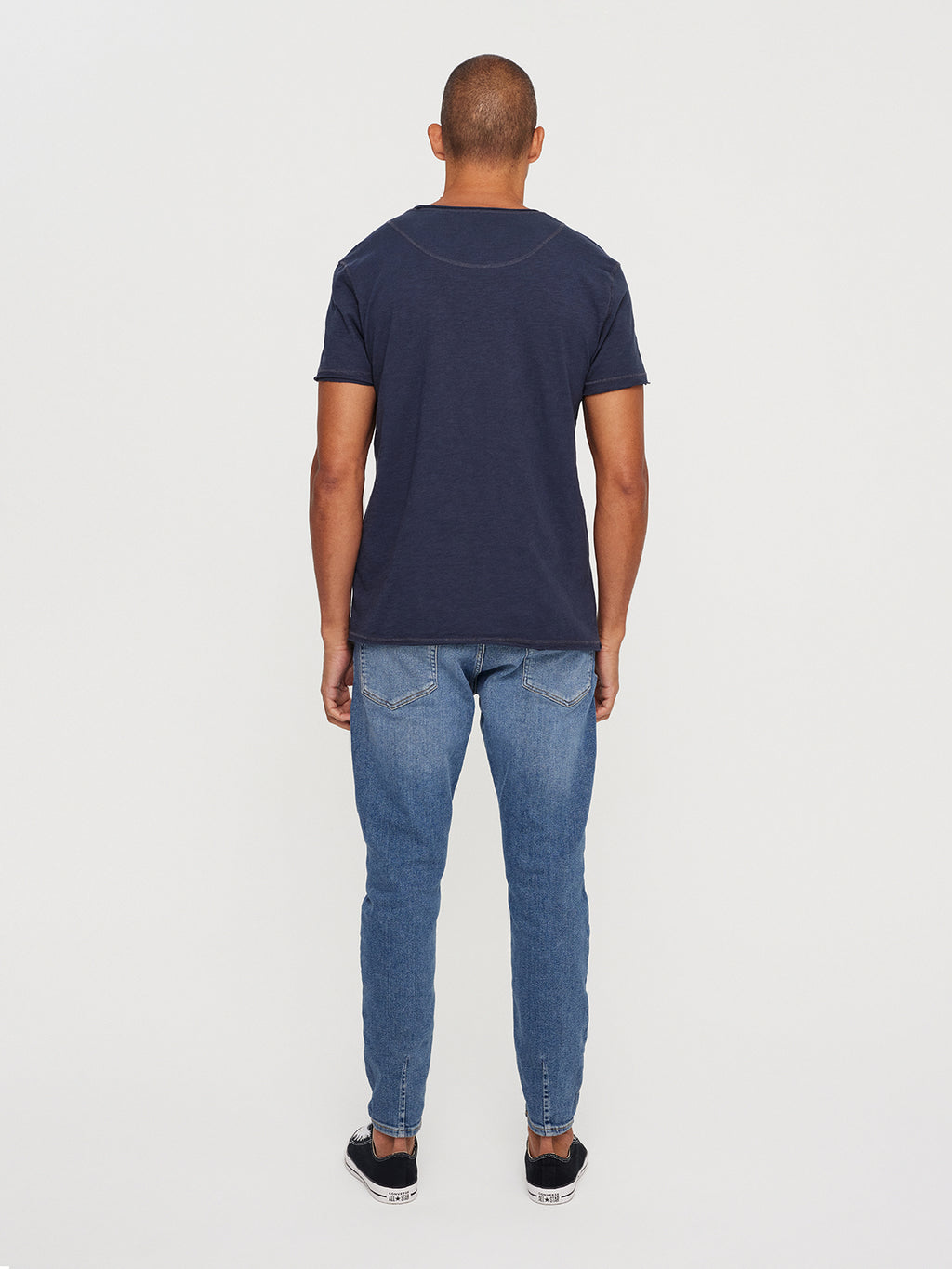 A person faces away from the camera, wearing GABBAs Konrad Straight Slub Tee in navy with blue jeans and black sneakers against a white backdrop.