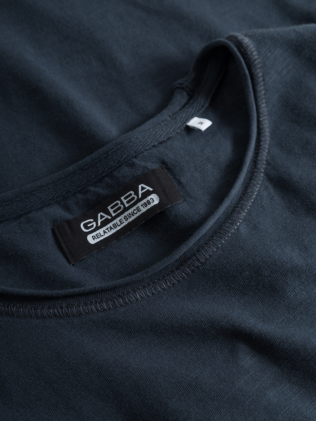 Close-up of a navy T-shirt featuring a tag that reads GABBA RELIABLE SINCE 1983, showcasing its casual neckline stitching. A partially visible size S tag is tucked in. Made from soft cotton slub yarn, the Konrad Straight Slub Tee offers style and comfort.