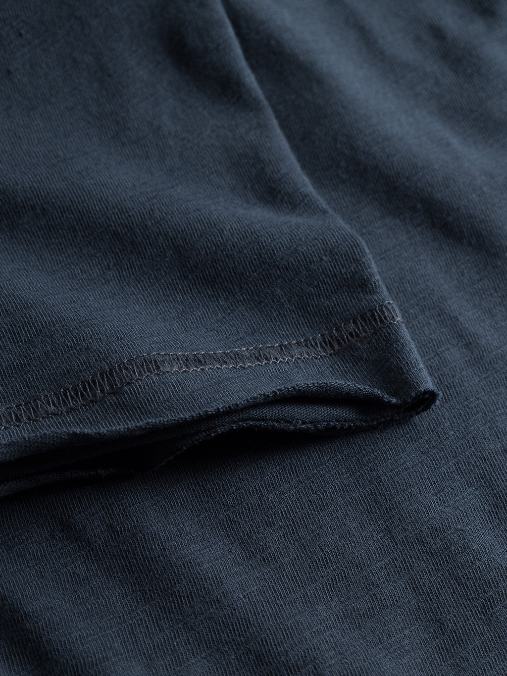 Close-up of GABBAs Konrad Straight Slub Tee in Navy, showcasing a textured surface from cotton slub yarn. The fabric has a neat hemline with precise stitching, creating a smooth yet slightly wavy finish, making it soft and perfect for a casual look.