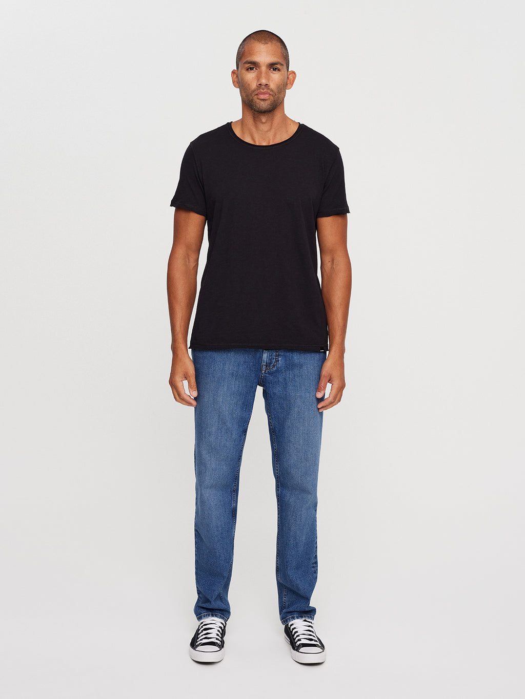 A man stands against a white background, wearing the GABBA Konrad Straight Slub Tee in black, paired with blue jeans and black sneakers with white soles. His arms are relaxed at his sides as he looks straight ahead in this regular fit ensemble.