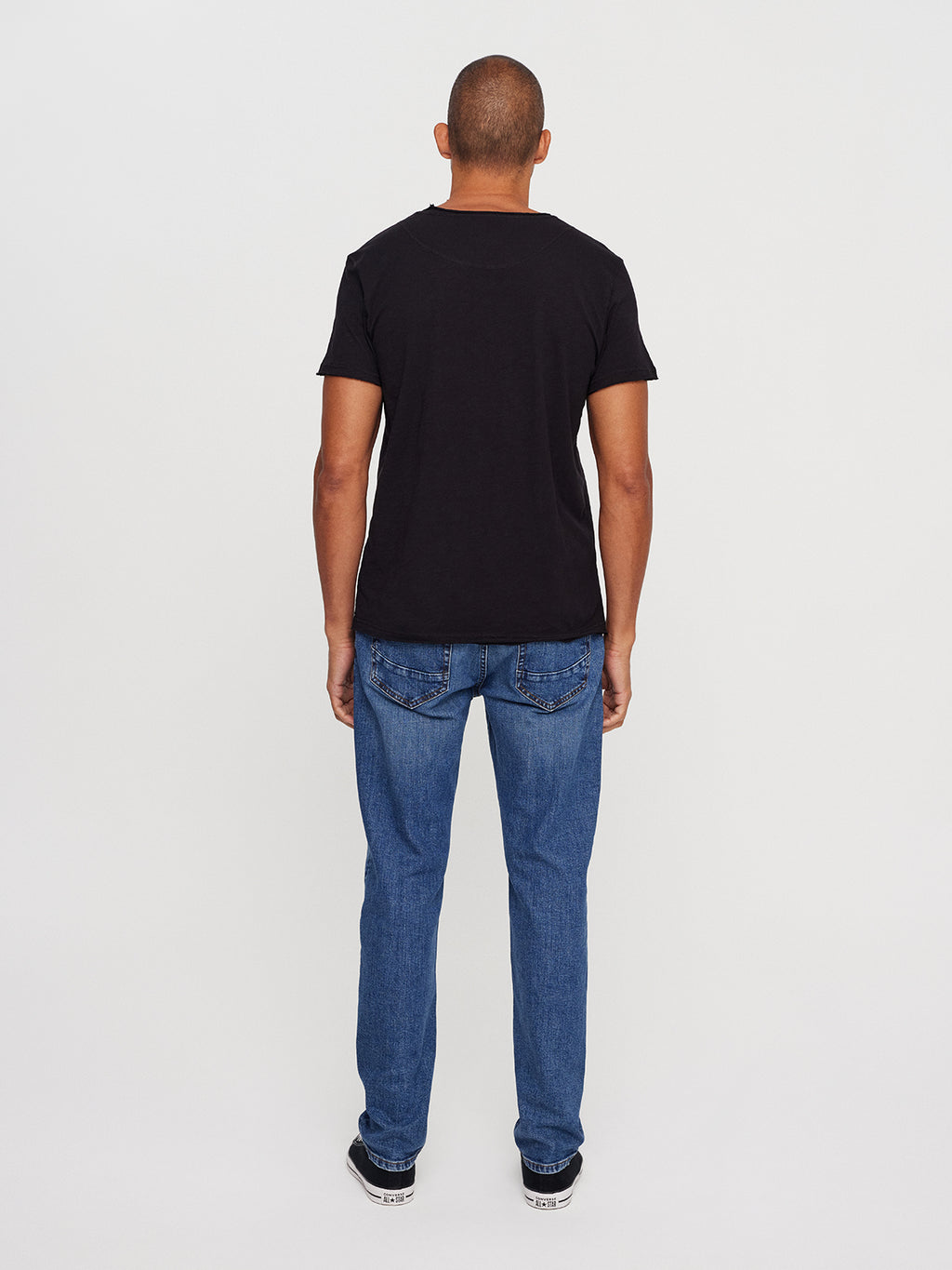 A man in a GABBA Konrad Straight Slub Tee - Black, with blue jeans and black sneakers, stands facing away against a white background.