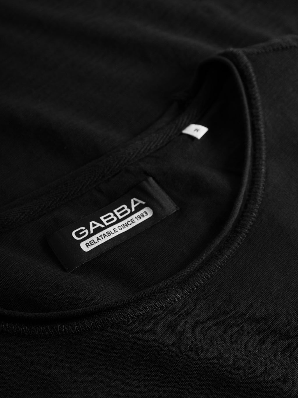 Close-up of the black Konrad Straight Slub Tee from GABBA, crafted from cotton slub yarn. The neatly folded regular-fit shirt features a label reading Relatable Since 1983 and a small white size tag.