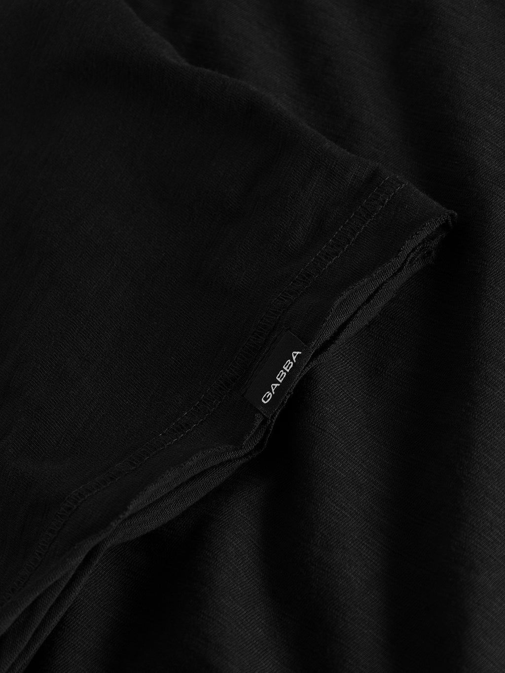 Close-up of the Konrad Straight Slub Tee in black from GABBA, featuring a small black tag with GABBA in white. The textured surface hints at cotton slub yarn, indicating its a regular fit basic tee.