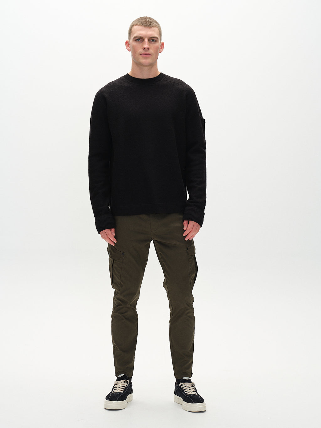 A person stands against a white background wearing a black long-sleeve sweater, Pisa Cargo K3280 Dale Pants - Army by GABBA, and black sneakers with white soles. The slim tapered fit of the olive green cargo pants with side pockets enhances the sleek look.