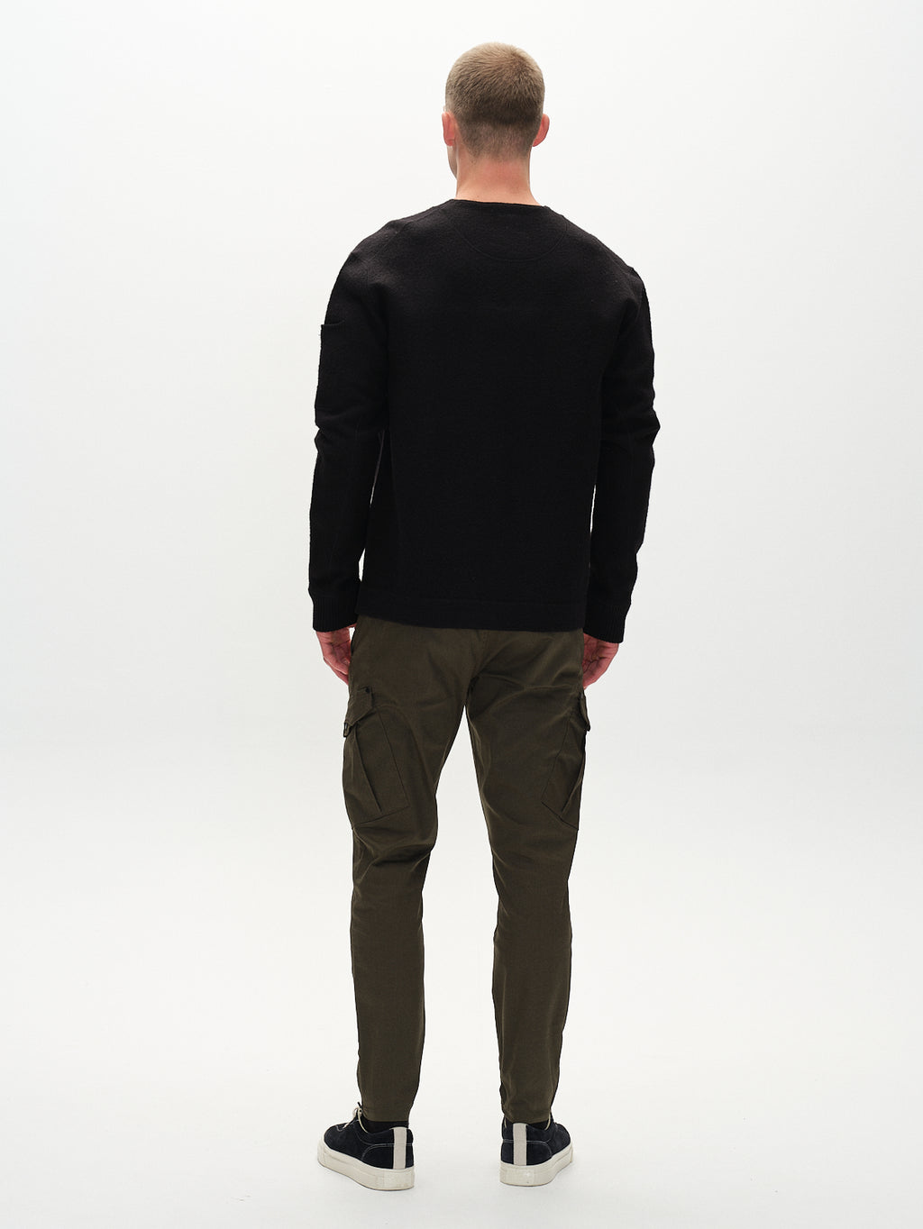A man wears a black sweater and GABBAs Pisa Cargo K3280 Dale Pants in Army. Hes facing away in a minimalist, neutral-toned setting, where the pants functional side pockets and slim tapered fit are enhanced by his black sneakers.