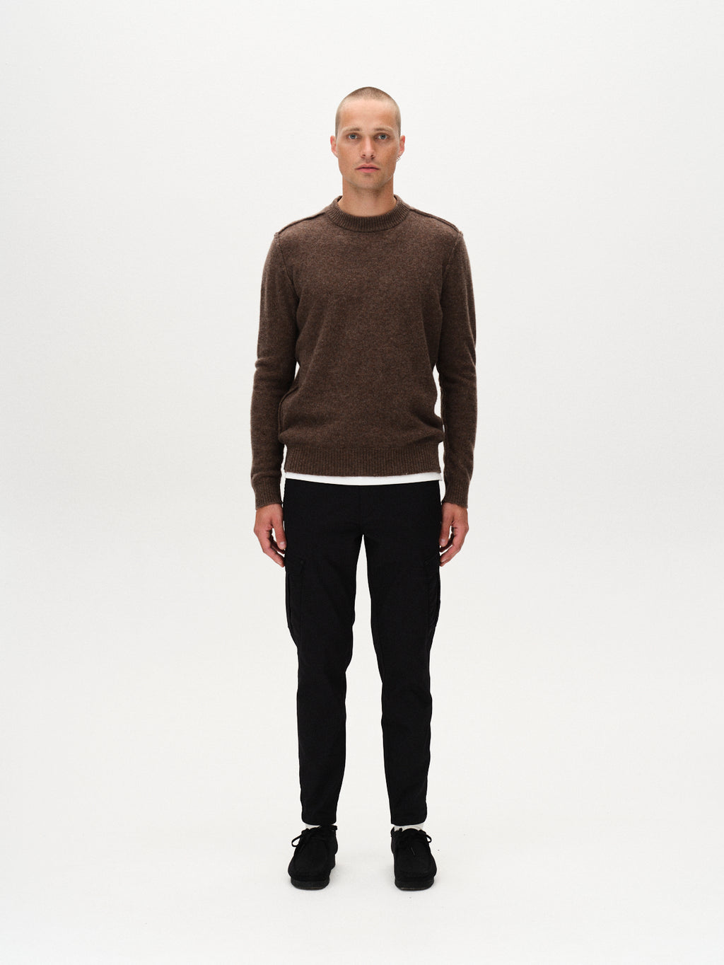 A person with short hair stands facing forward against a plain white background, wearing a brown sweater, black shoes, and GABBA Pisa Cargo K3280 Dale Pants in black with a slim tapered fit made from super stretchy fabric.