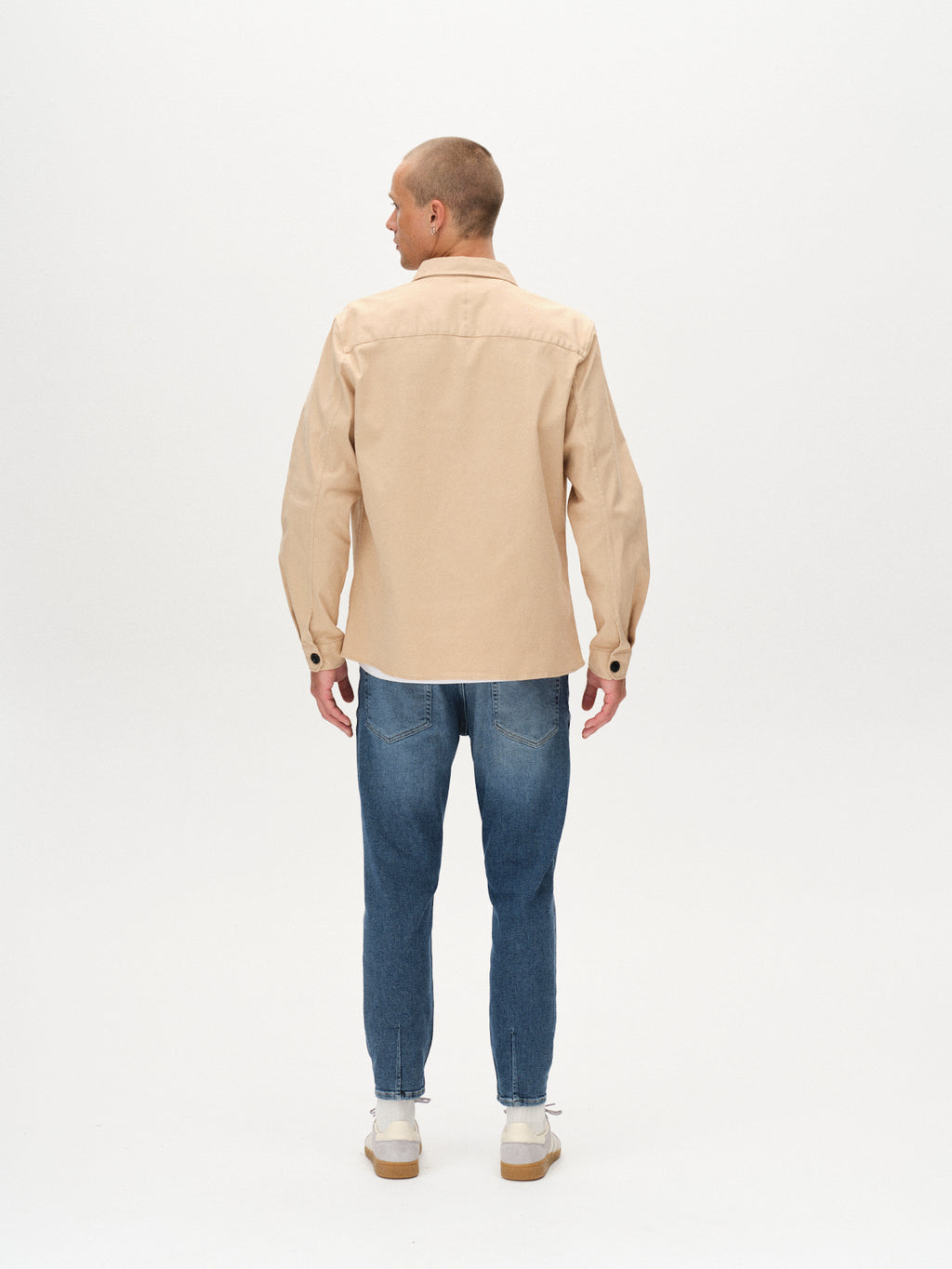 A man with a shaved head faces away, wearing the GABBA Topper LS Shirt in Humus layered over a shirt, paired with blue jeans and white sneakers, set against a plain white background.