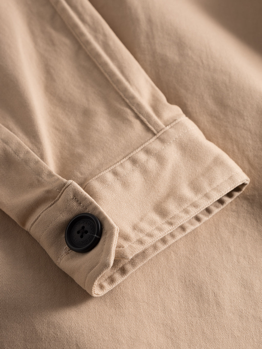Close-up of the Topper LS Shirt - Humus by GABBA, showcasing its beige cuff, black button, fabric texture, and stitch details. Ideal for layering as an overshirt, it adds sophistication to any ensemble.