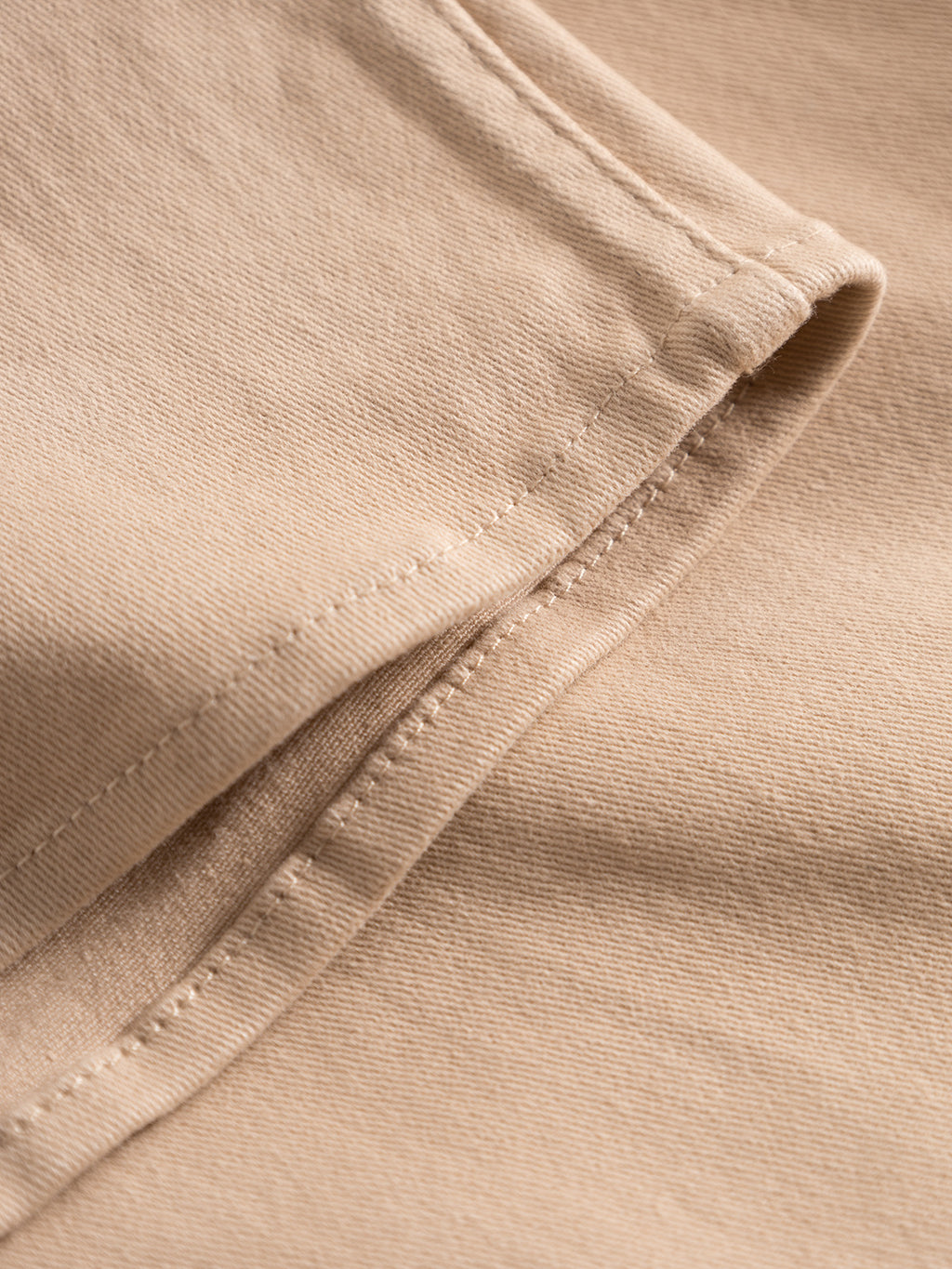 The GABBA Topper LS Shirt - Humus features a close-up of beige fabric with visible stitching and hem, offering a soft, smooth texture similar to an overshirt. Ideal for layering, it showcases detail with its chest pockets.