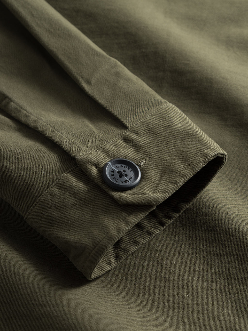 Close-up of the GABBA Topper LS Shirt - Army fabric, highlighting detailed stitching and texture. Features a buttoned cuff with engraved gray buttons that subtly contrast with the fabrics matte olive green tone.