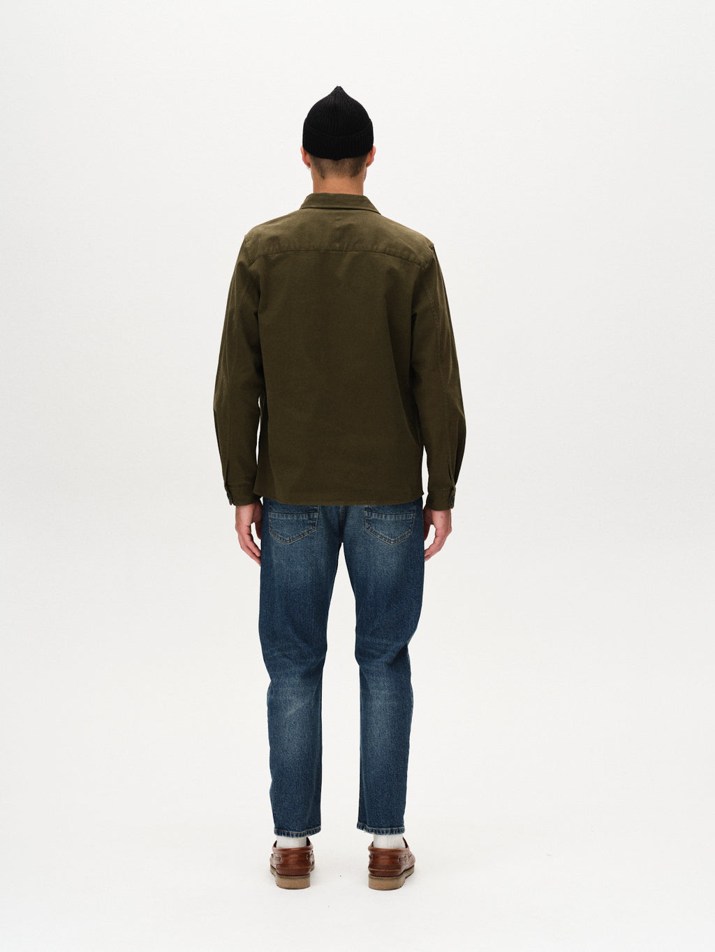A person wearing a black beanie, GABBA Topper LS Shirt - Army, blue jeans, and brown shoes stands facing away against a plain white background.