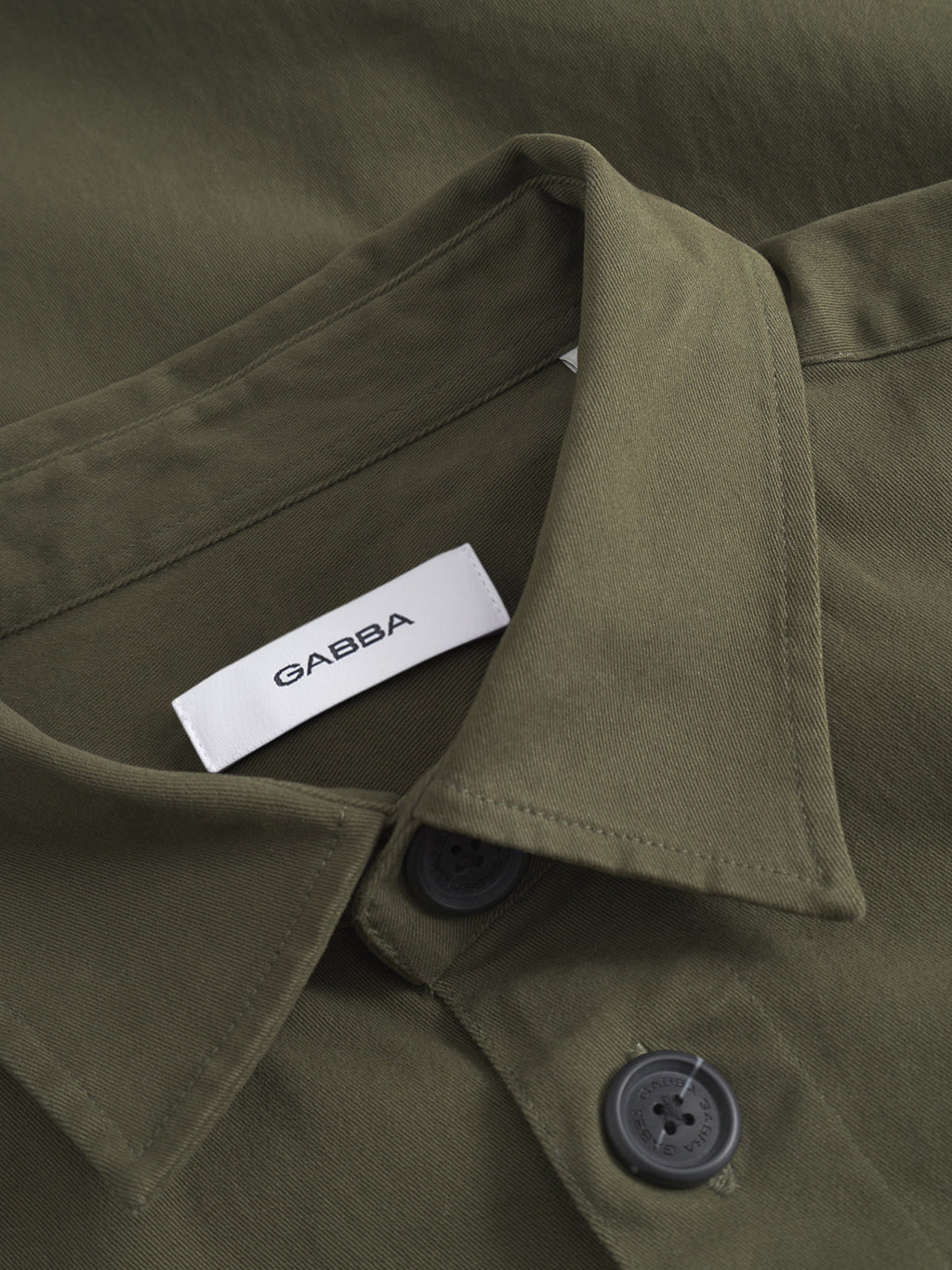 Close-up of the GABBA Topper LS Shirt - Army, an olive green overshirt featuring black button closure and a white inside collar label with the GABBA brand name.