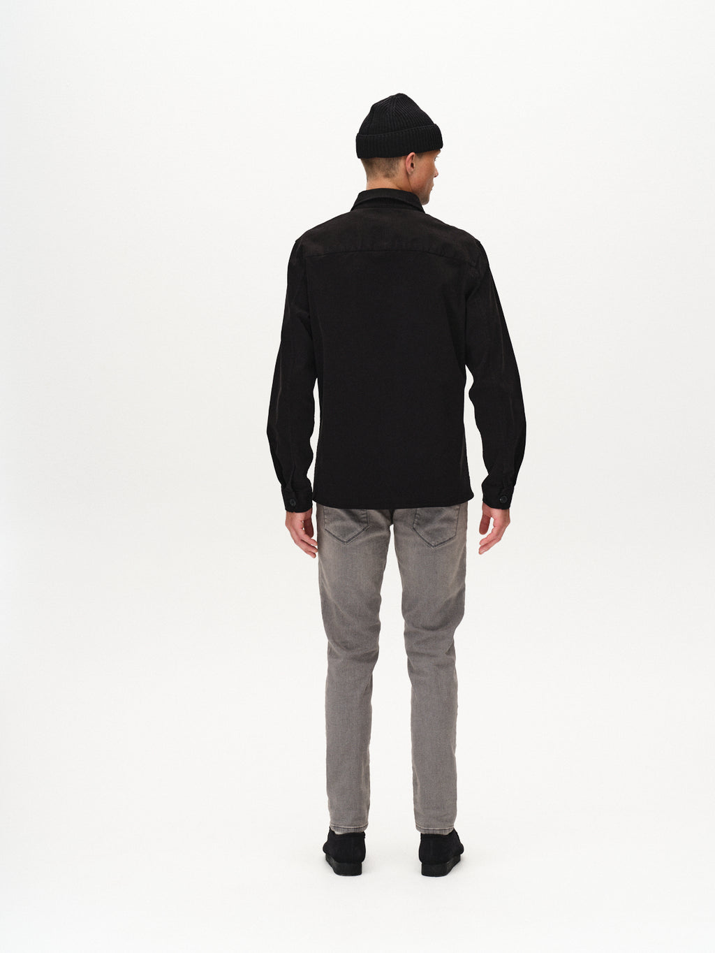A man stands facing away from the camera on a white background, wearing the GABBA Topper LS Shirt in black, paired with light gray jeans, black shoes, and a black beanie.