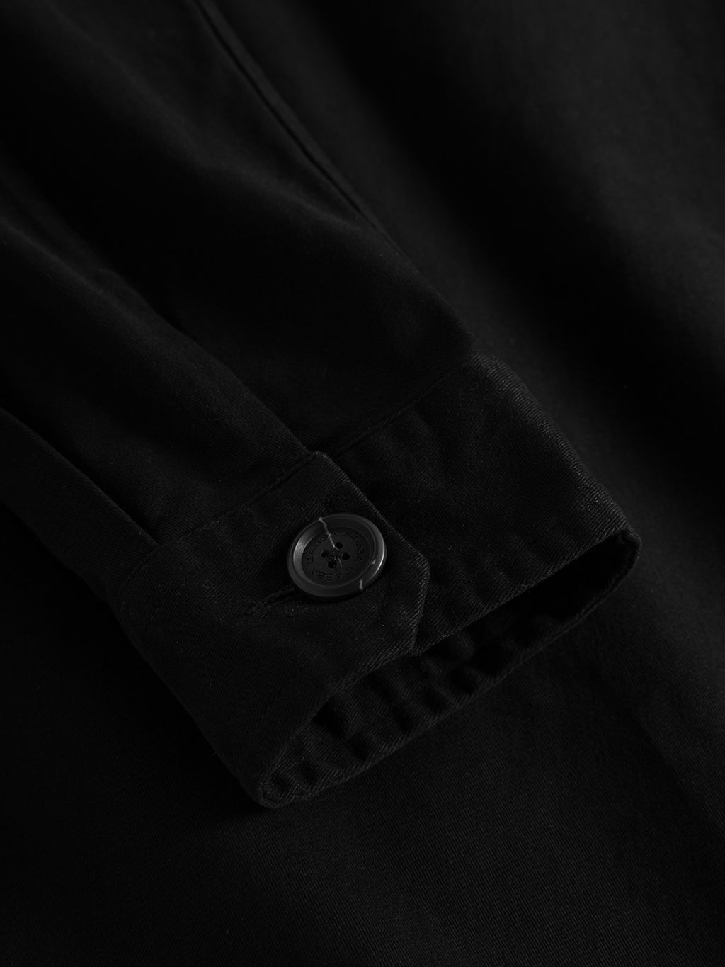 A close-up of the Topper LS Shirt - Black by GABBA showcases its sleek black fabric and a buttoned cuff, ideal for stylish layering. The finely stitched material highlights the button and thread against a slightly crumpled backdrop, perfect for an effortlessly stylish overshirt look.