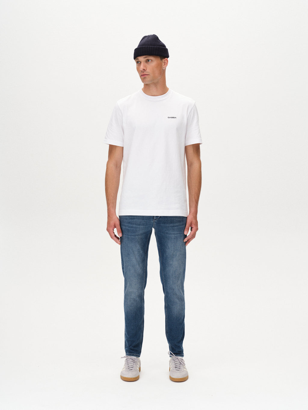 A man wearing a black beanie, white t-shirt, GABBA Rey K3866 TENCEL™ Jeans in denim wash, and white sneakers stands against a plain white background, looking slightly left with his arms relaxed for a laid-back vibe.