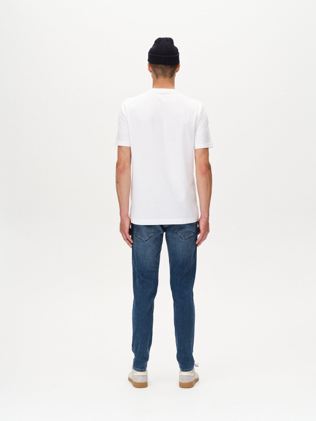 A person stands facing away from the camera in a black beanie, white T-shirt, GABBA Rey K3866 TENCEL™ Jeans - Denim wash, and white sneakers—creating a relaxed look against a plain white background.