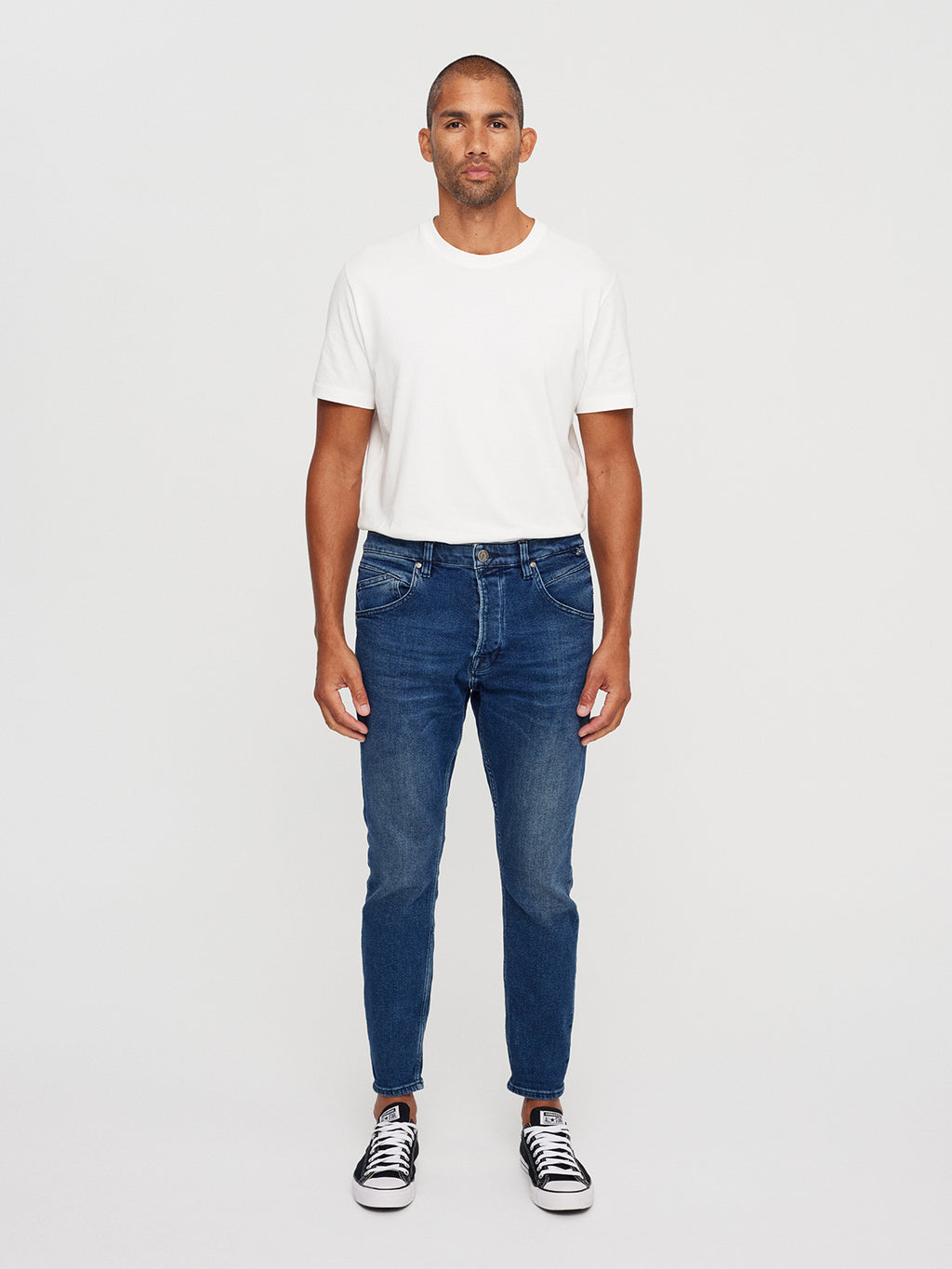 A man with short hair stands against a plain white background, wearing a white t-shirt, GABBAs Alex K3868 Jeans in denim wash, and black sneakers. His casual style is evident from his relaxed posture and arms resting by his sides.