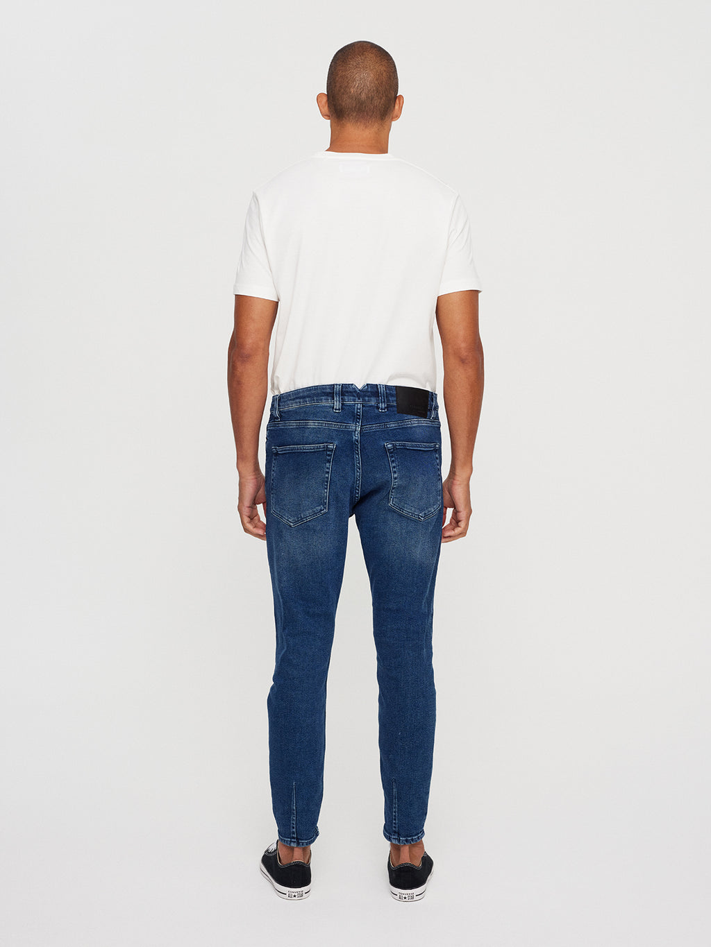 A person with their back to the camera showcases a trend-focused look. Theyre wearing a white t-shirt, GABBAs Alex K3868 Jeans - Denim wash, and black sneakers, with a phone tucked into their jeans waistband against a plain white background.