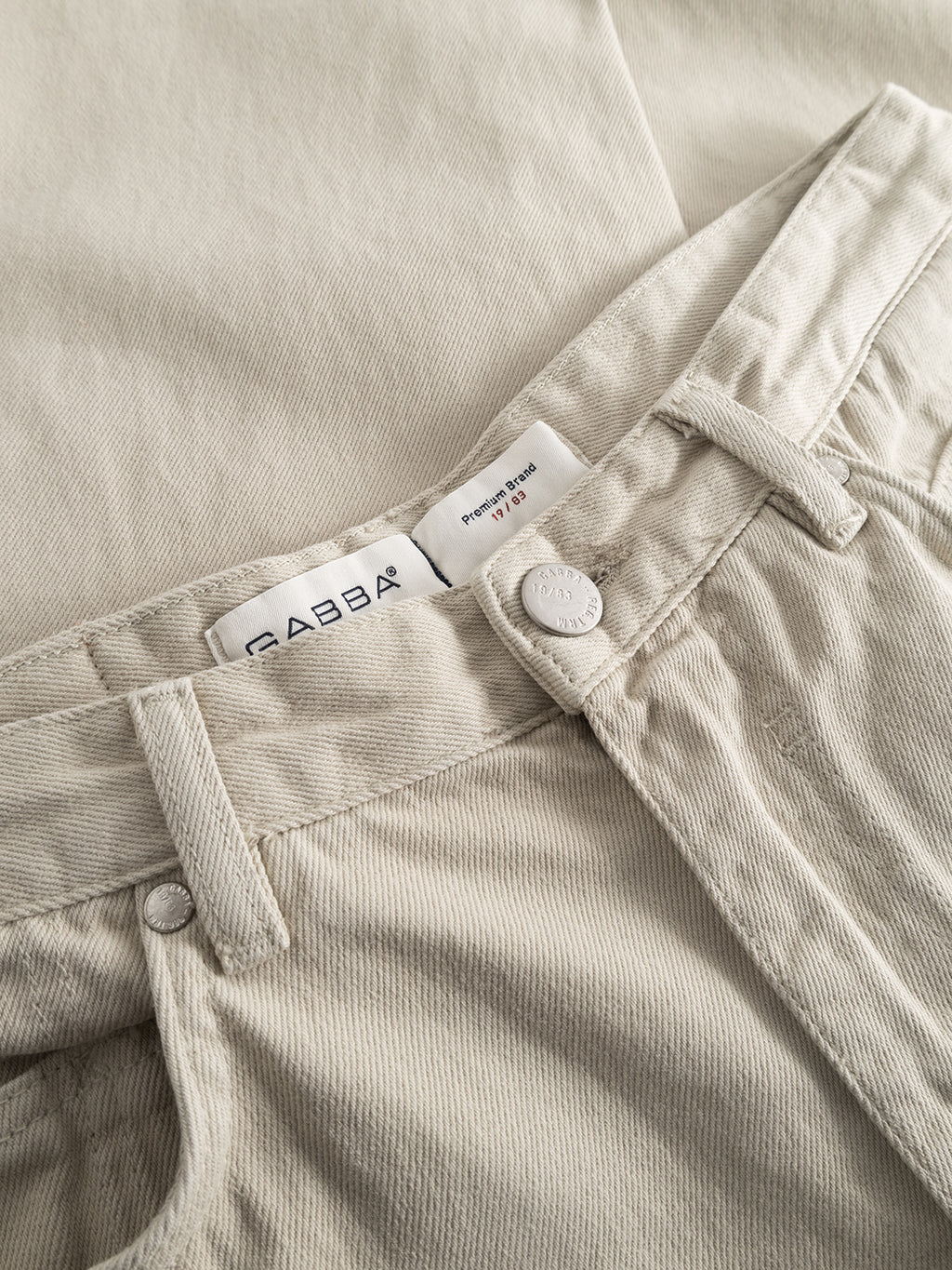 A close-up of the beige Zem K3992 Jeans by GABBA exudes a vintage flair. The focus is on the waistband and button, with a visible inner label reading GABBA. The fabrics smooth texture channels urban 90s style, with the jeans neatly arranged.