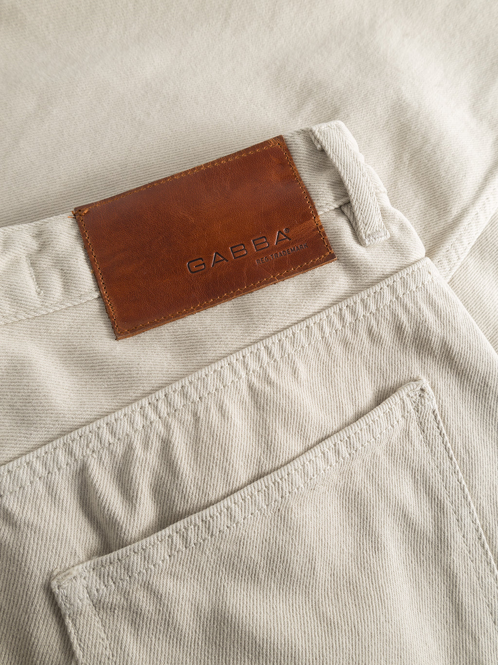 Close-up of GABBAs Zem K3992 Jeans in beige, showcasing a brown leather patch with the brand name embossed. The image captures the textured fabric and a back pocket beneath the patch, enhancing the urban 90’s vibe with its tapered leg design.