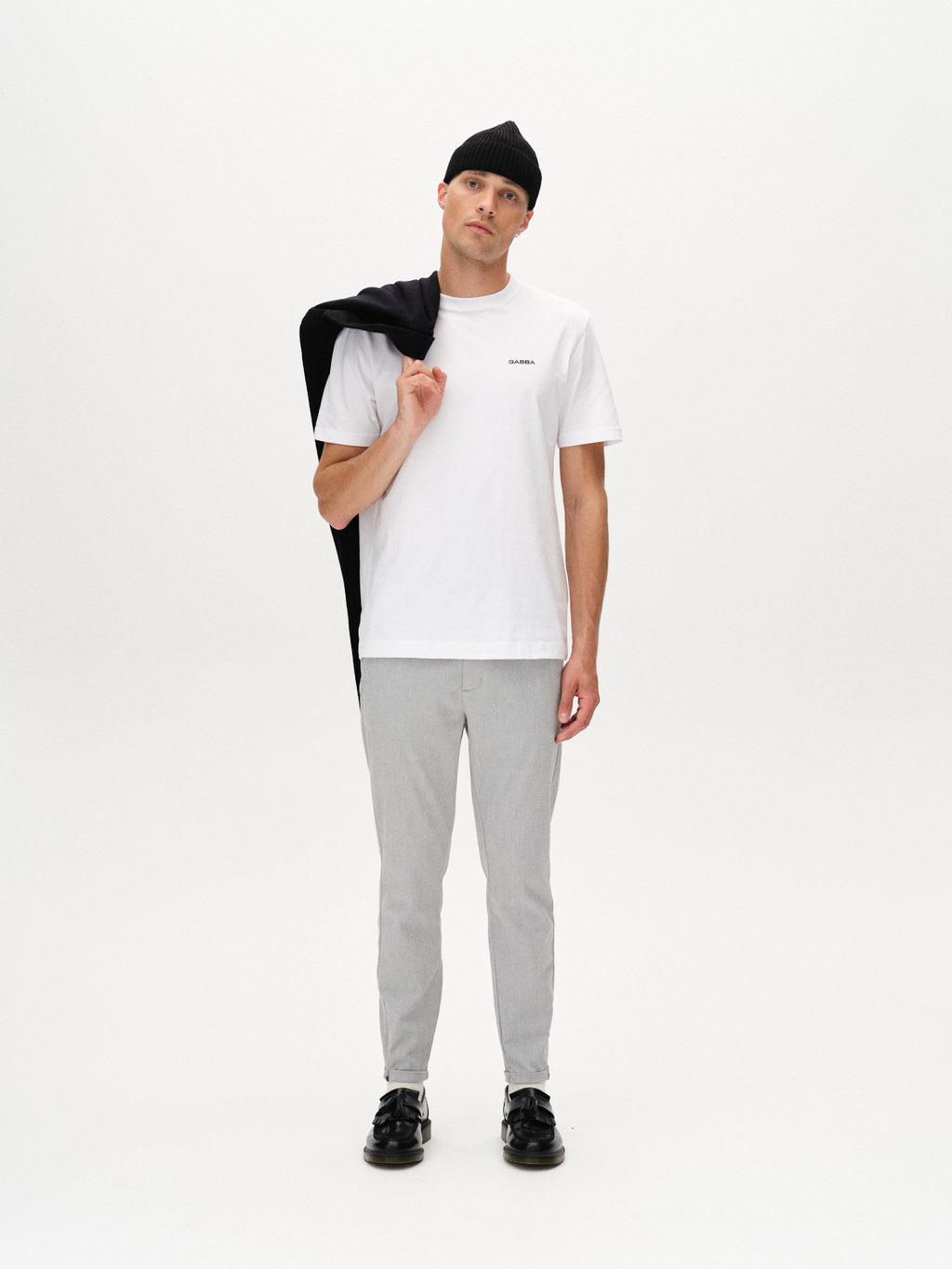 A man in a black beanie, white t-shirt, and Gabba Pisa Domo Pants - Monument stands against a white background, holding a black jacket over his shoulder for a sleek metro style look.