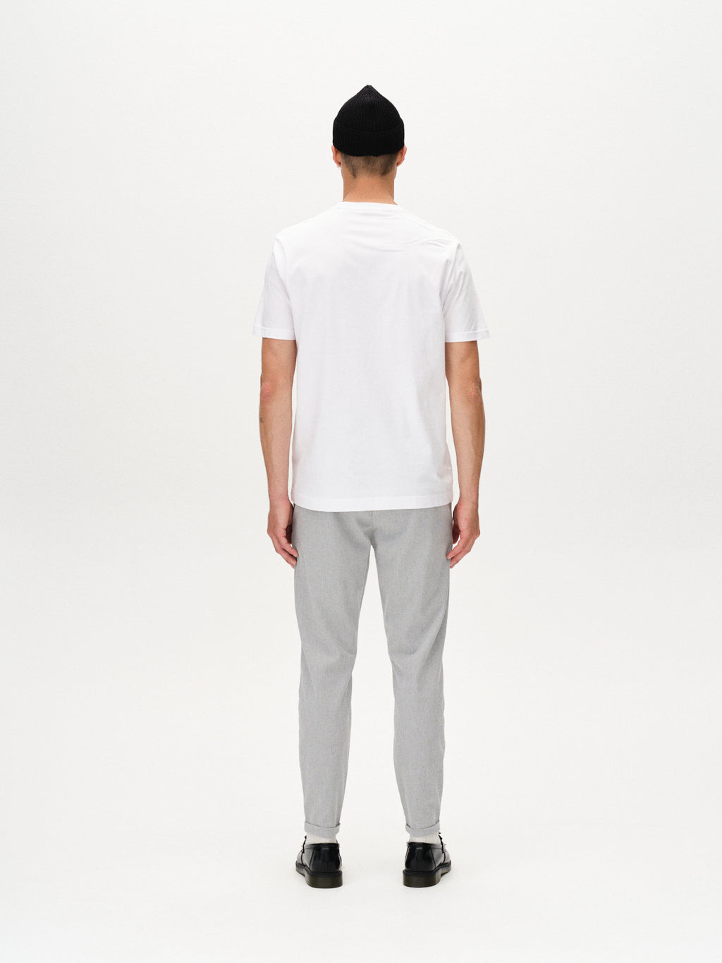 A person stands with their back to the camera, wearing a white t-shirt, Gabba Pisa Domo Pants - Monument in light gray high-stretch cotton-blend, black shoes, and a black beanie against a plain white background.