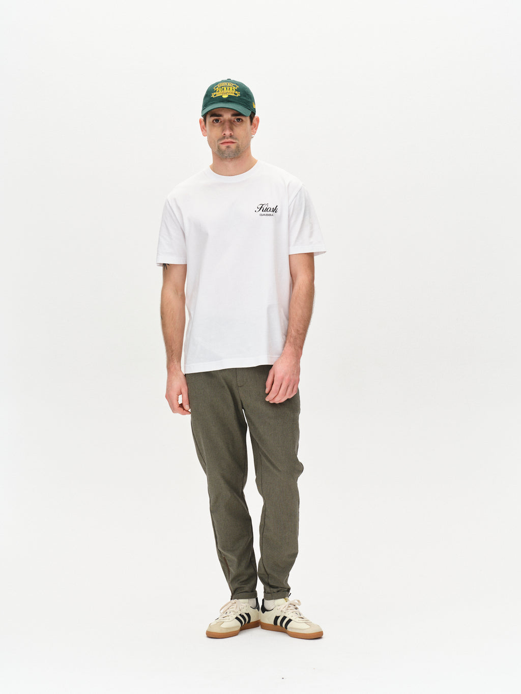 A man stands against a plain white background in metro style, wearing a green baseball cap, a printed white T-shirt, Gabba Pisa Domo Pants - SS25Army, and white sneakers with black stripes.