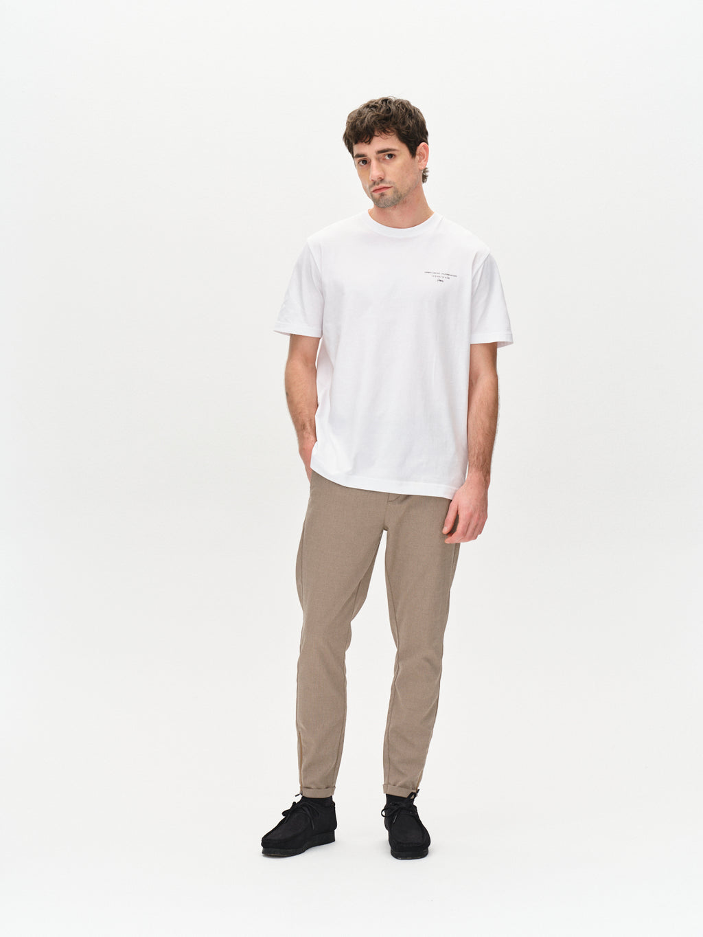 A man with short brown hair and a neutral expression stands against a white background wearing Gabba Pisa Domo Pants in khaki, a metro style white T-shirt, and black shoes. One hand rests by his side; the other is slightly bent.