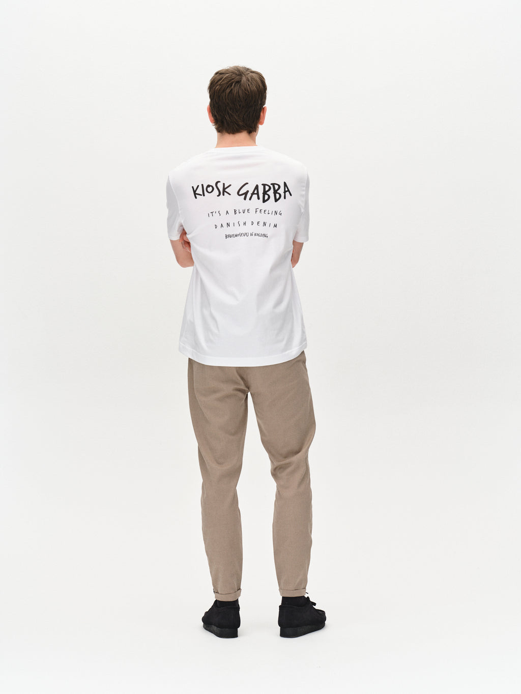 A person faces away, wearing a white KIOSK GABBA T-shirt with back text, Pisa Domo Pants - Khaki by Gabba in high-stretch cotton blend, and black shoes, styled metro against a plain white background.