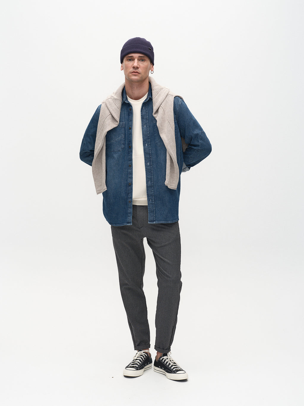 A man stands against a plain white background, sporting metro style with a dark beanie, denim button-up over a white top, grey sweater draped over his shoulders, Gabba Pisa Domo Pants in Dark Grey, and black sneakers. His hands rest behind his back.