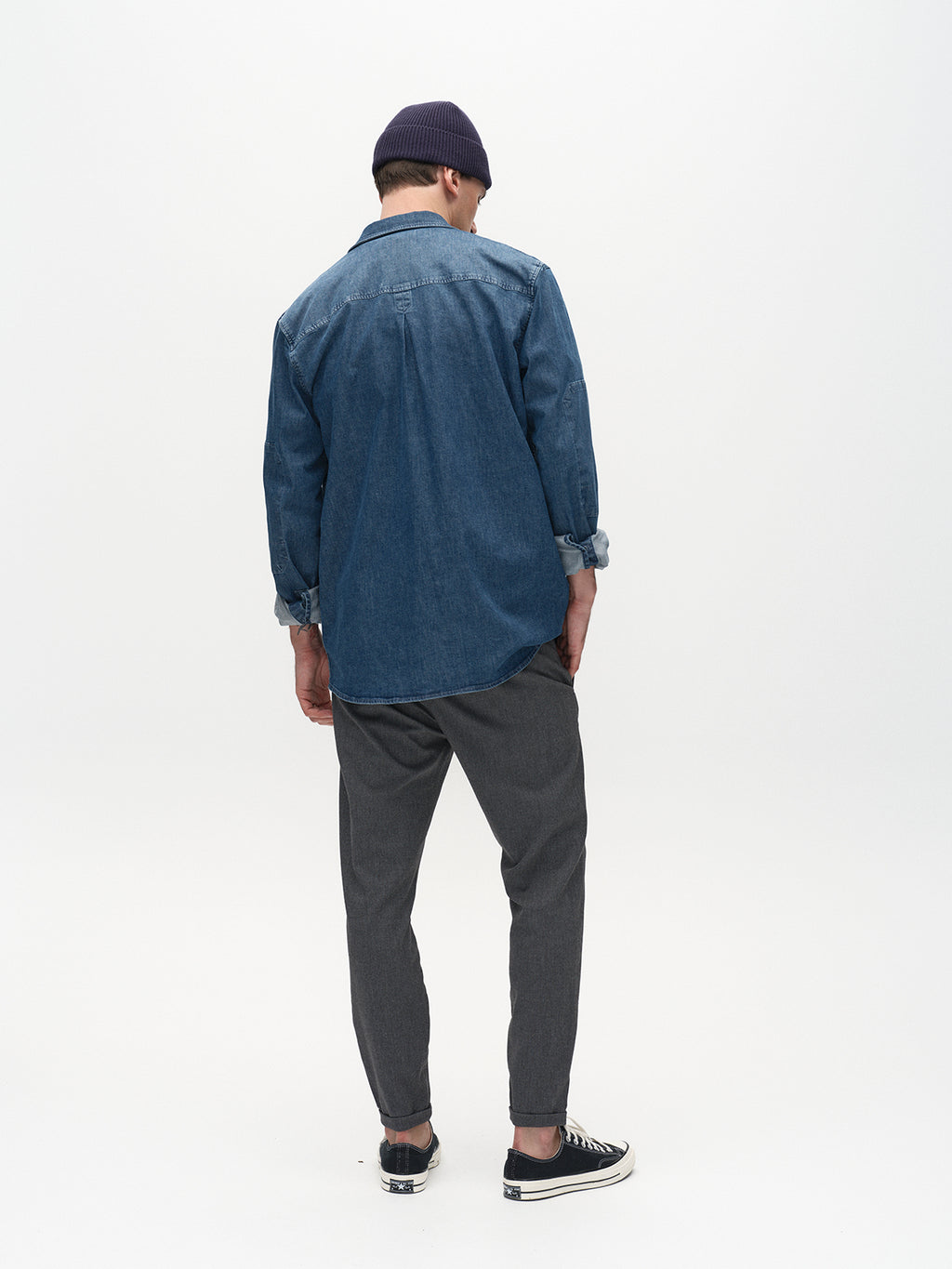 A person stands facing away in a metro style, wearing a blue denim shirt, Gabba Pisa Domo Pants in dark grey, black sneakers, and a dark beanie against a plain white background.
