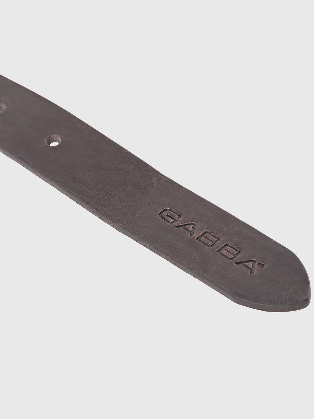 Close-up of the Okay Belt - Brown from GABBA, made of premium dark brown leather featuring the word GABBA embossed. The design includes two visible holes and tapers to a rounded end against a light gray background.