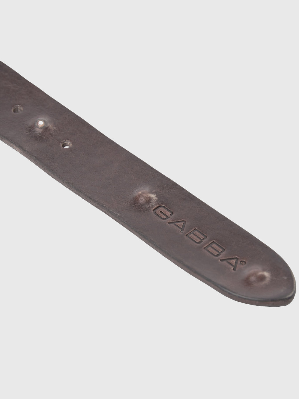 Close-up of the Okay Point Belt by GABBA in dark brown leather, featuring three small fastening holes and embossed with the brand name on the end. Set against a light gray background.