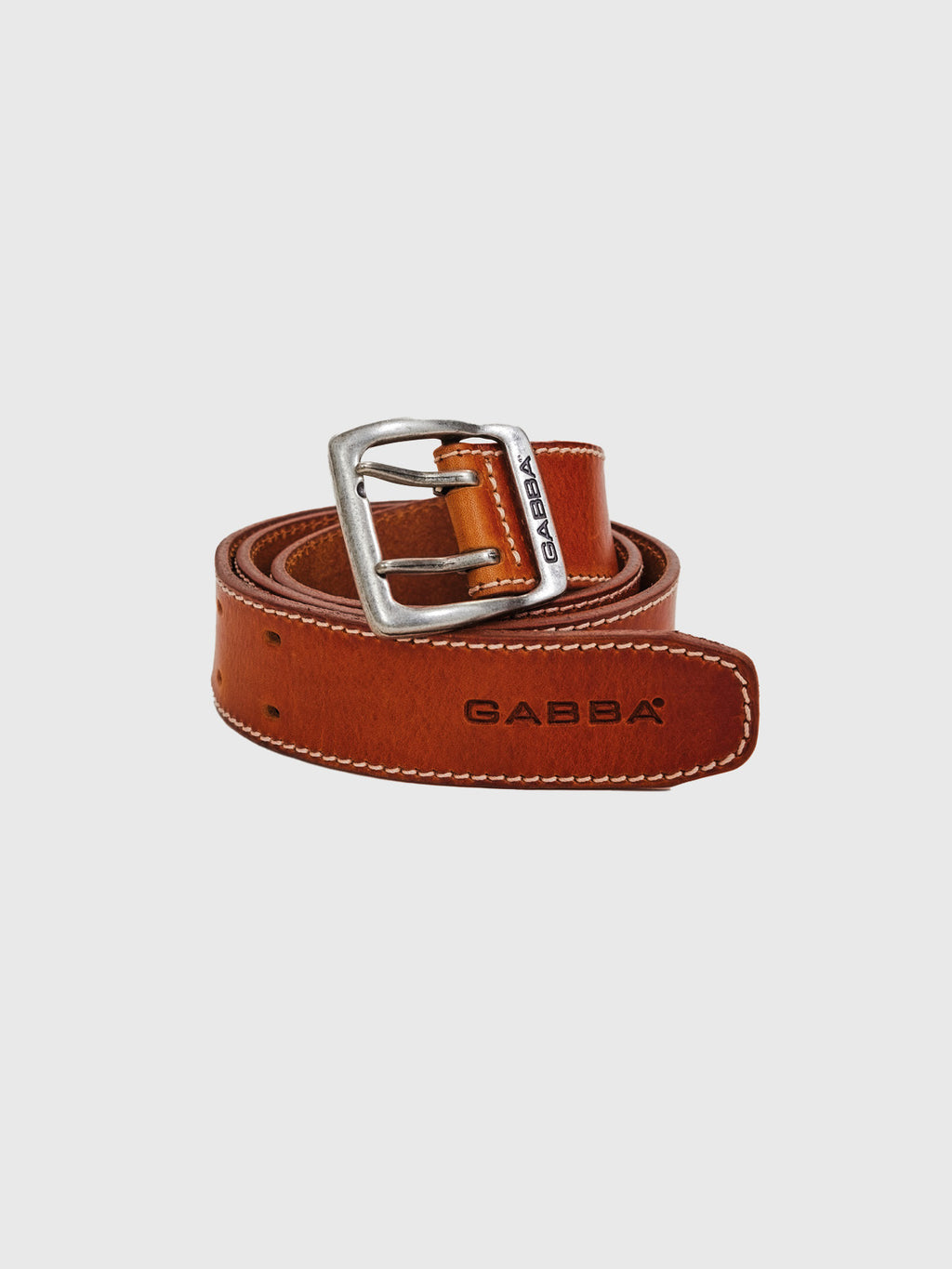 The Basta Stitch Belt in cognac, by GABBA, is made from high-quality leather with white stitching along the edges. It features a silver buckle and has GABBA embossed near the tip and engraved on the buckle. It lies coiled on a light gray background.
