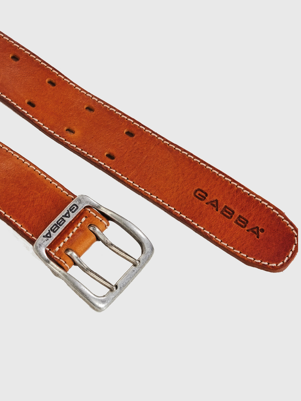 The Basta Stitch Belt - Cognac by GABBA features a timeless design with high-quality leather. The brown belt has white stitching, a silver GABBA-stamped buckle on the left, multiple holes on the right, and the iconic GABBA logo printed in black.