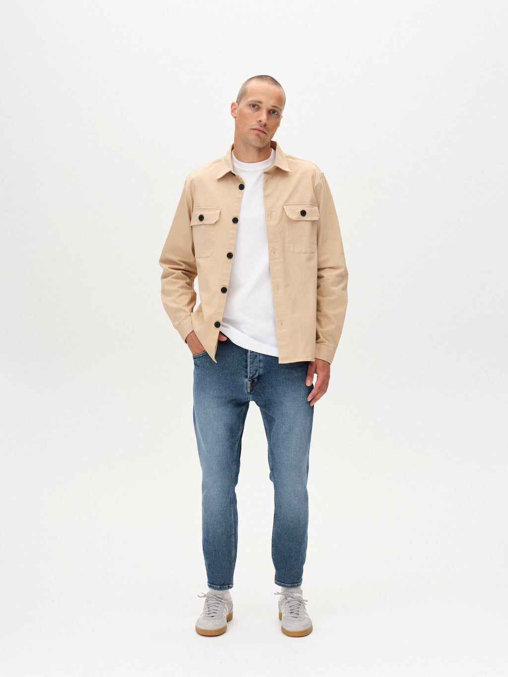 A man stands against a white background wearing GABBAs Alex K3948 Jeans in denim wash, paired with a beige button-up over a white t-shirt and light gray sneakers—an effortless look with one hand in his pocket and a neutral expression.
