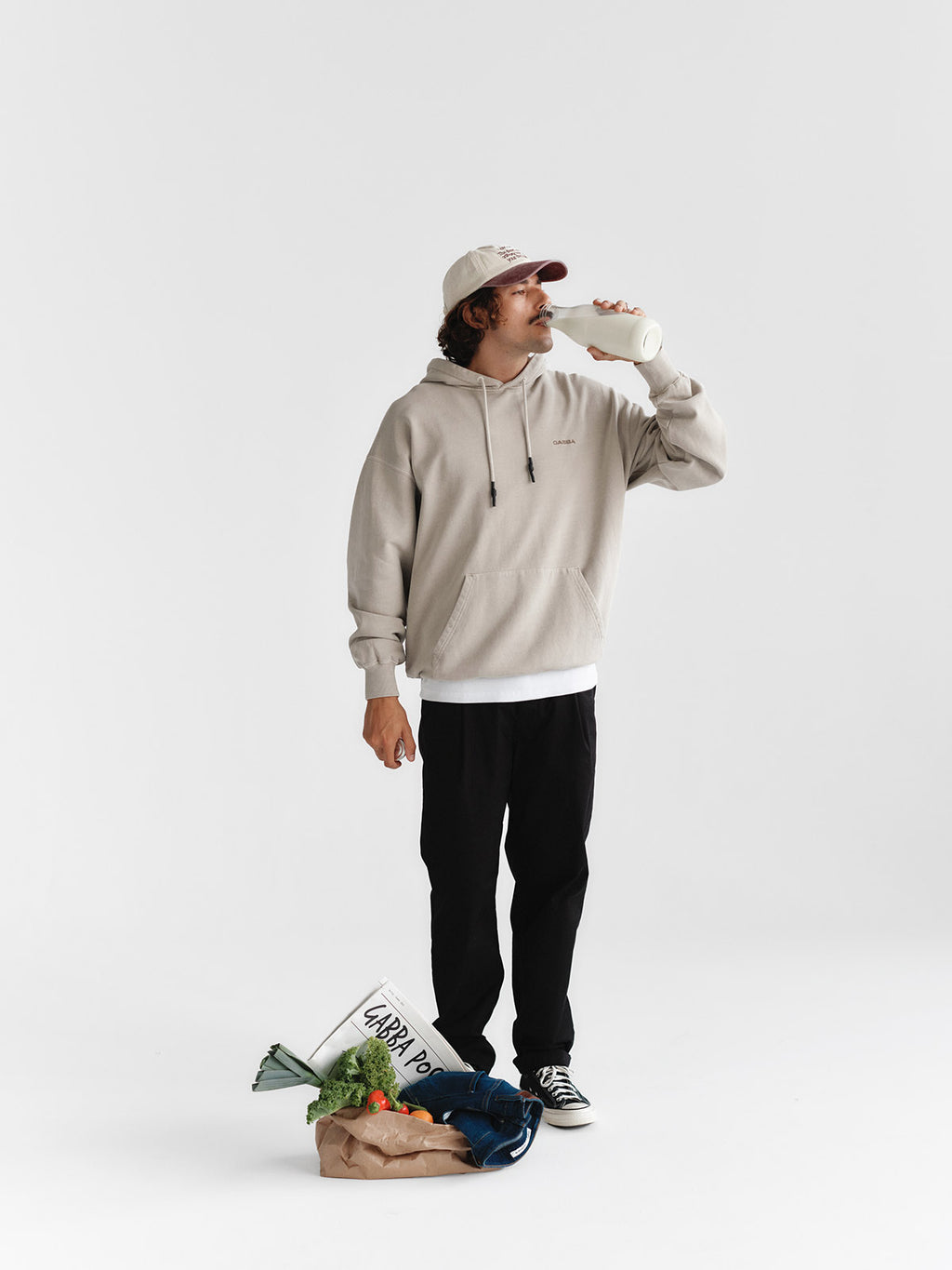 A man wears the Gabba Joe Unite - Black relaxed fit, tapered-leg pants paired with a beige hoodie and cap, sipping from a bottle. A grocery bag with veggies, baguette, and newspaper rests at his feet against a plain white background.