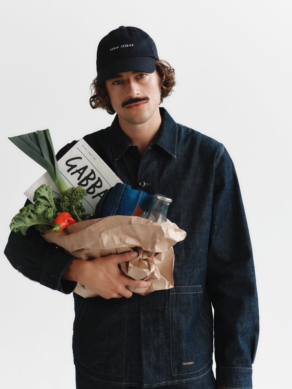 A mustached man in Gabbas Jap Beam dark blue denim jacket and a black cap holds a brown grocery bag with vegetables and bread labeled Gabba against a plain white background.