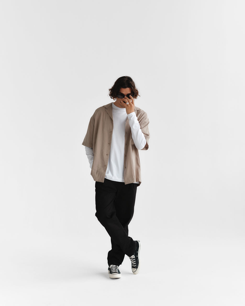 A person with wavy hair wearing sunglasses, a Gabba Jens Over Resort Yari SS shirt in Sand Greige over a long-sleeved white tee, black pants, and sneakers stands against a white background, leaning slightly with one hand near their face.