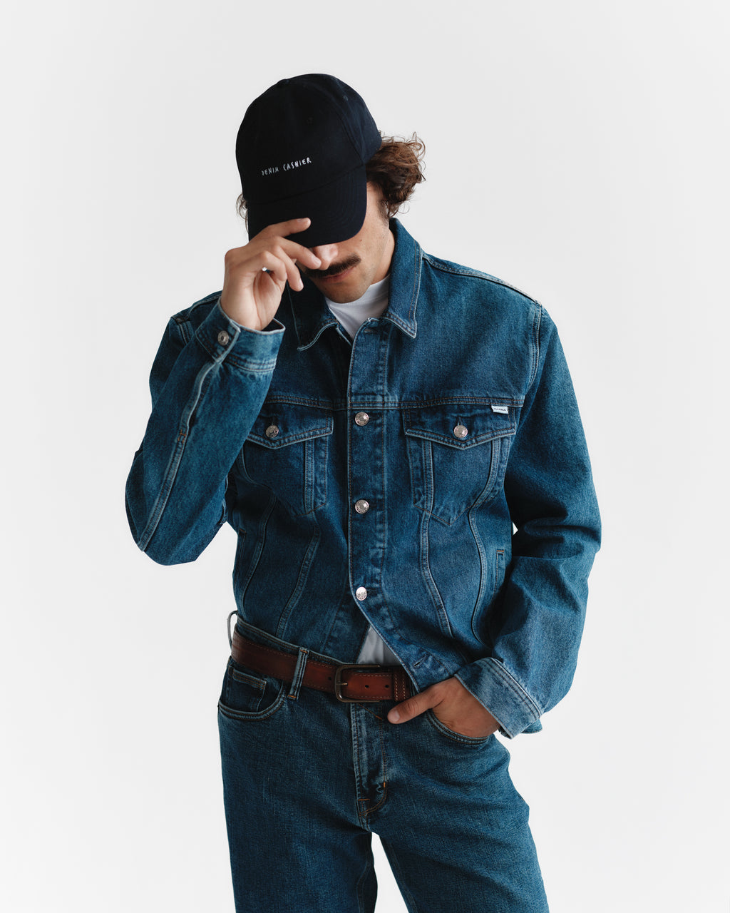 A person clad in a denim jacket and jeans adjusts a Gabba Cashier Cap - Night Sky, partially covering their face with the brim. The plain white background highlights their casual, stylish look.