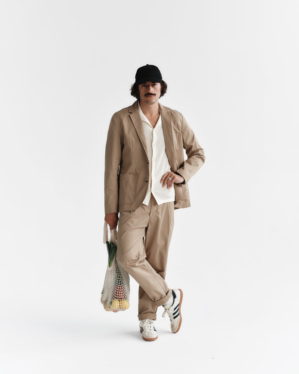 Wearing Gabbas Joe Unite - Sand suit with tapered leg pants and a white shirt, a person stands against a white background in a black cap and sneakers, holding a net bag filled with fruit to complete the casual look.