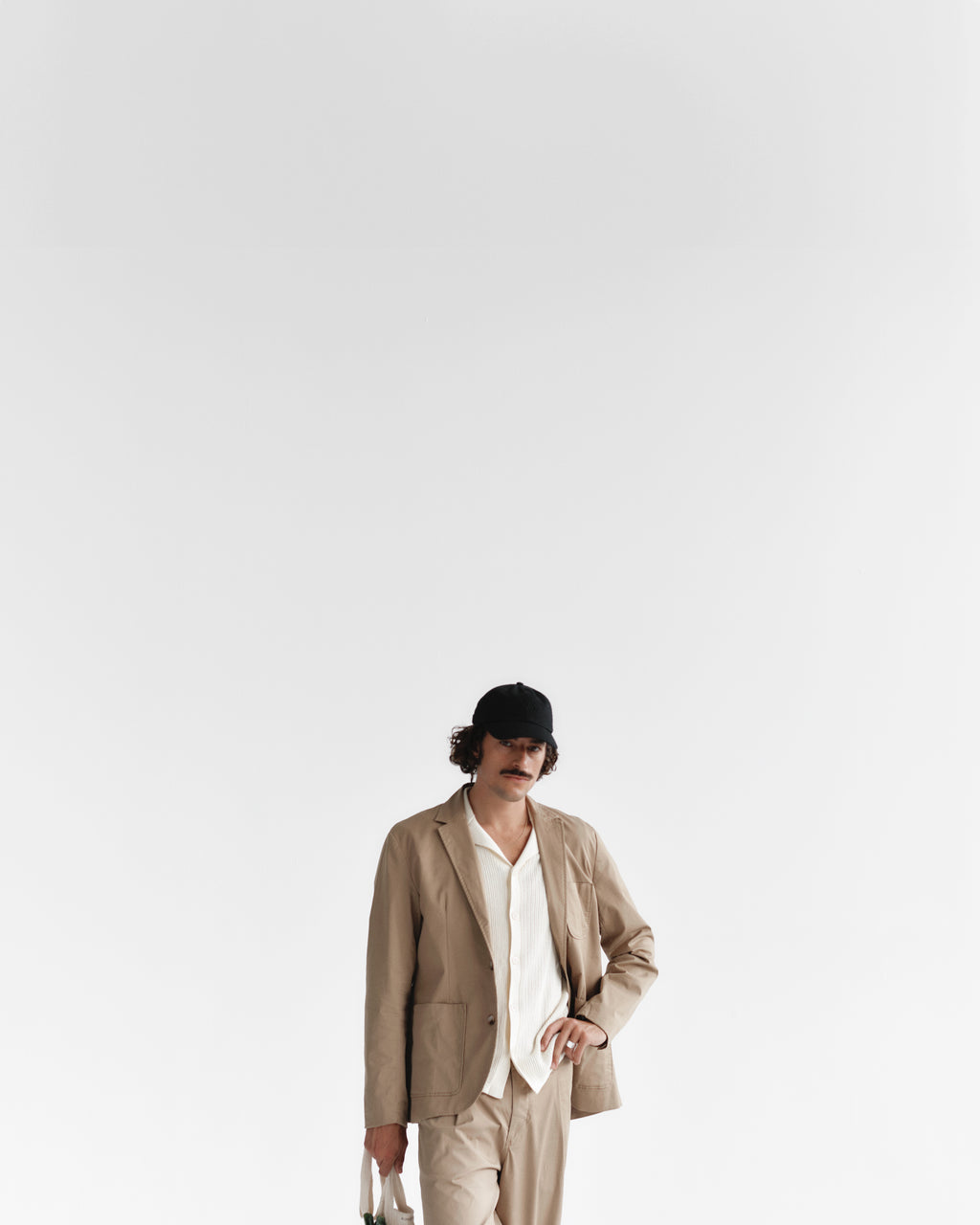 A person with curly hair, wearing a black cap and Gabbas Geno Resort SS - Ecru Light draped over a classic white shirt, stands holding a suitcase against a plain white background, exuding a retro vibe.