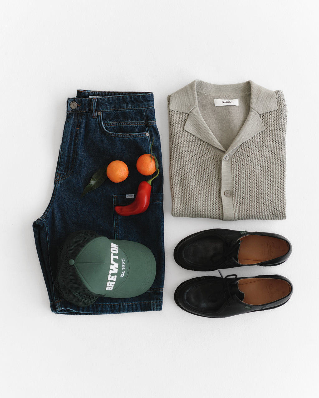 A neatly folded beige knitted shirt, Ugo Cargo Fone - Mid Blue Denim from Gabba, and black leather shoes are arranged on a white surface. Nearby are a green baseball cap, two oranges, and a red chili pepper.