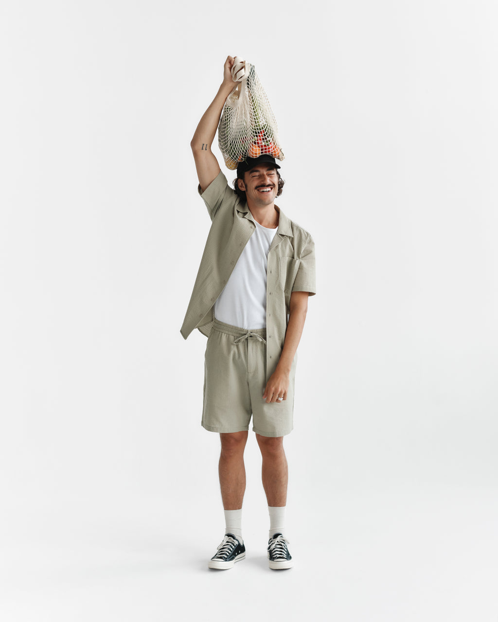 A smiling person in summer attire—wearing Gabba’s Tommi Seer - Green Sea shirt and sneakers—holds a mesh bag of produce above their head against a plain white background.