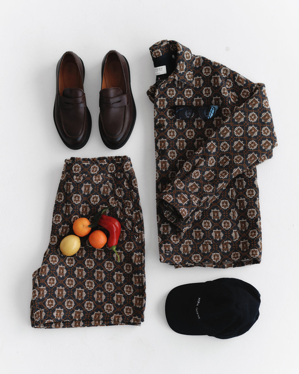 A stylish statement outfit from Gabba, the Fede Box Star - Multi Pattern brown jacket and pants set, is paired with black loafers and a matching cap. The loose-fitting ensemble is artfully displayed on a white surface with fruits like lemon, orange, and bell pepper placed on the pants.
