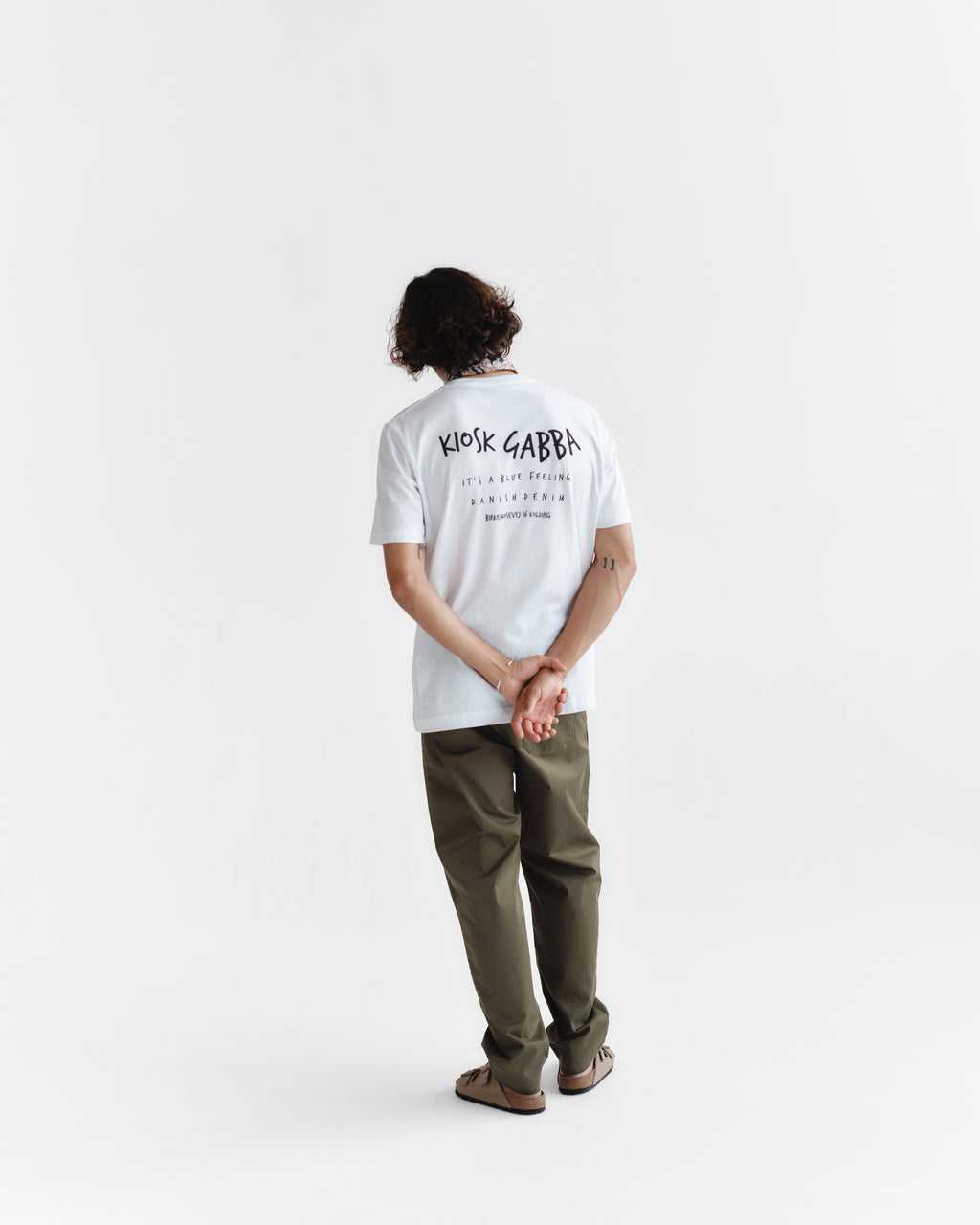 A person with wavy hair, facing away, wears a white t-shirt and Gabbas Joe Unite - Army relaxed-fit olive green pants. Their hands are clasped behind their back against a simple white backdrop, enhancing the comfortable casual look.