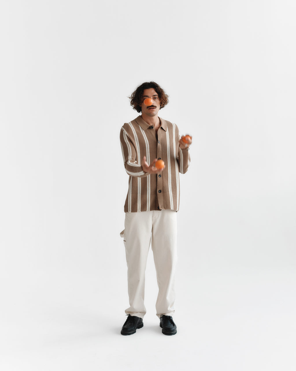 A person with curly hair, wearing Gabbas Dezz Stripes LS in brown stripe and light cotton pants, is skillfully juggling three oranges against a plain white background.
